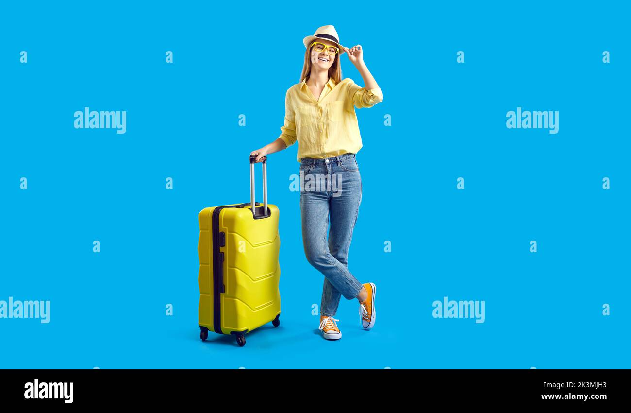 Portrait of happy female tourist standing with yellow suitcase on blue studio background Stock Photo