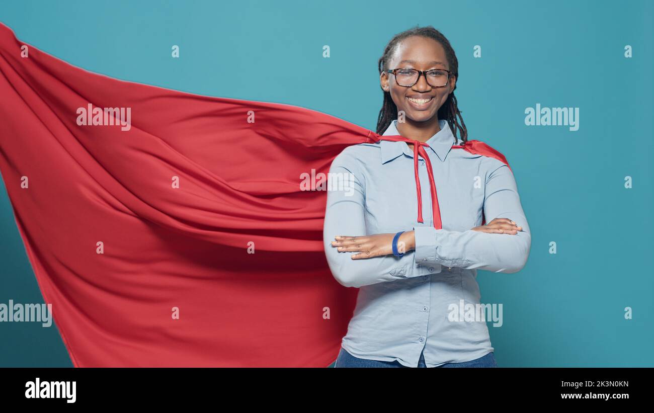 African american woman posing as superhero with red flying cape, wearing cartoon comic costume with cloak on camera. Feeling confident and powerful as hero, strong action character. Stock Photo