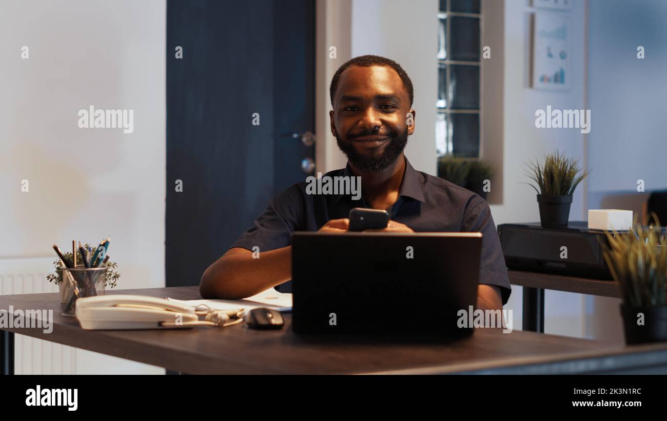 Portrait of smiling disruptive employee texting messages on smartphone, using internet app and working with online network on laptop. Planning executive marketing report late at night. Stock Photo