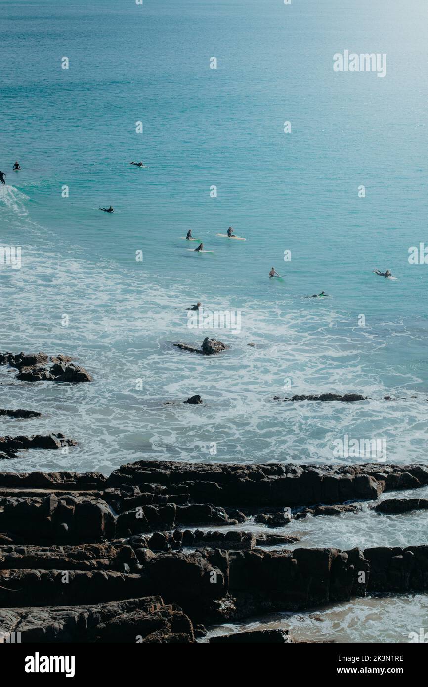 A vertical shot of people surfing in a blue sea against a rocky beach Stock Photo