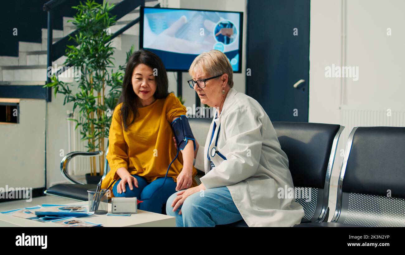 Elderly medic and asian woman doing hypertension measurement with tonometer at health center. Checking pulse pressure with cardiology test instrument, having medical appointment. Stock Photo