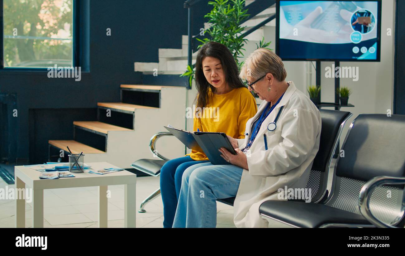 Elderly doctor and patient doing checkup visit in facility lobby, talking about treatment and medication. Taking notes on tablet and filling in medical report form to receive medical insurance. Stock Photo