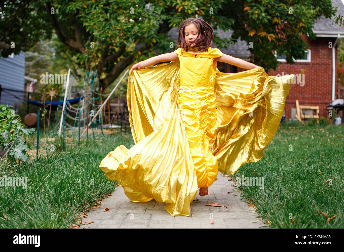 A girl in gold dress and wings leaps barefoot into air in backyard Stock Photo