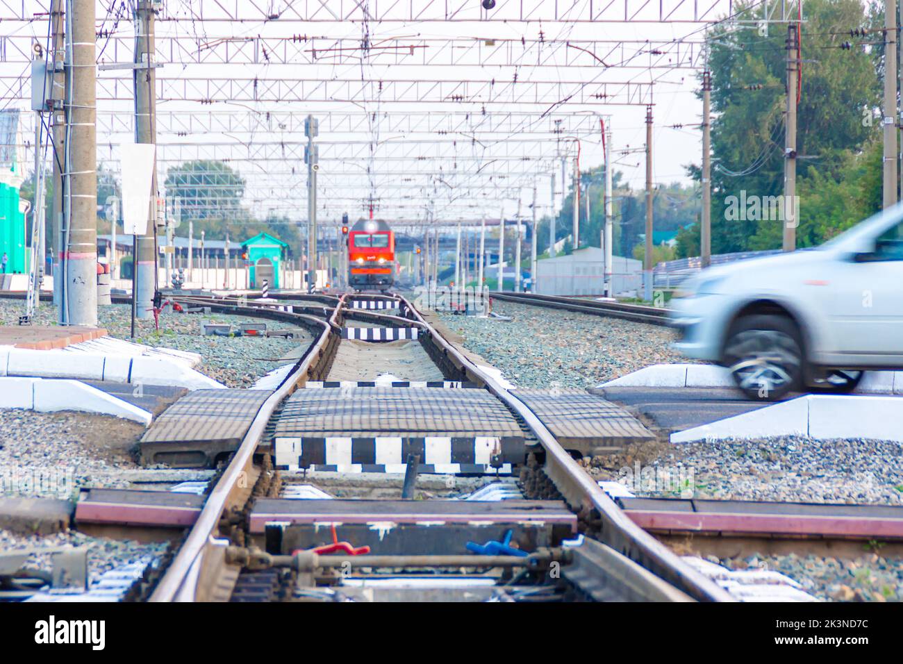 A red locomotive of Russian railways in the area of a small station is approaching a railway crossing that is dangerously crossed by a passenger car, Stock Photo