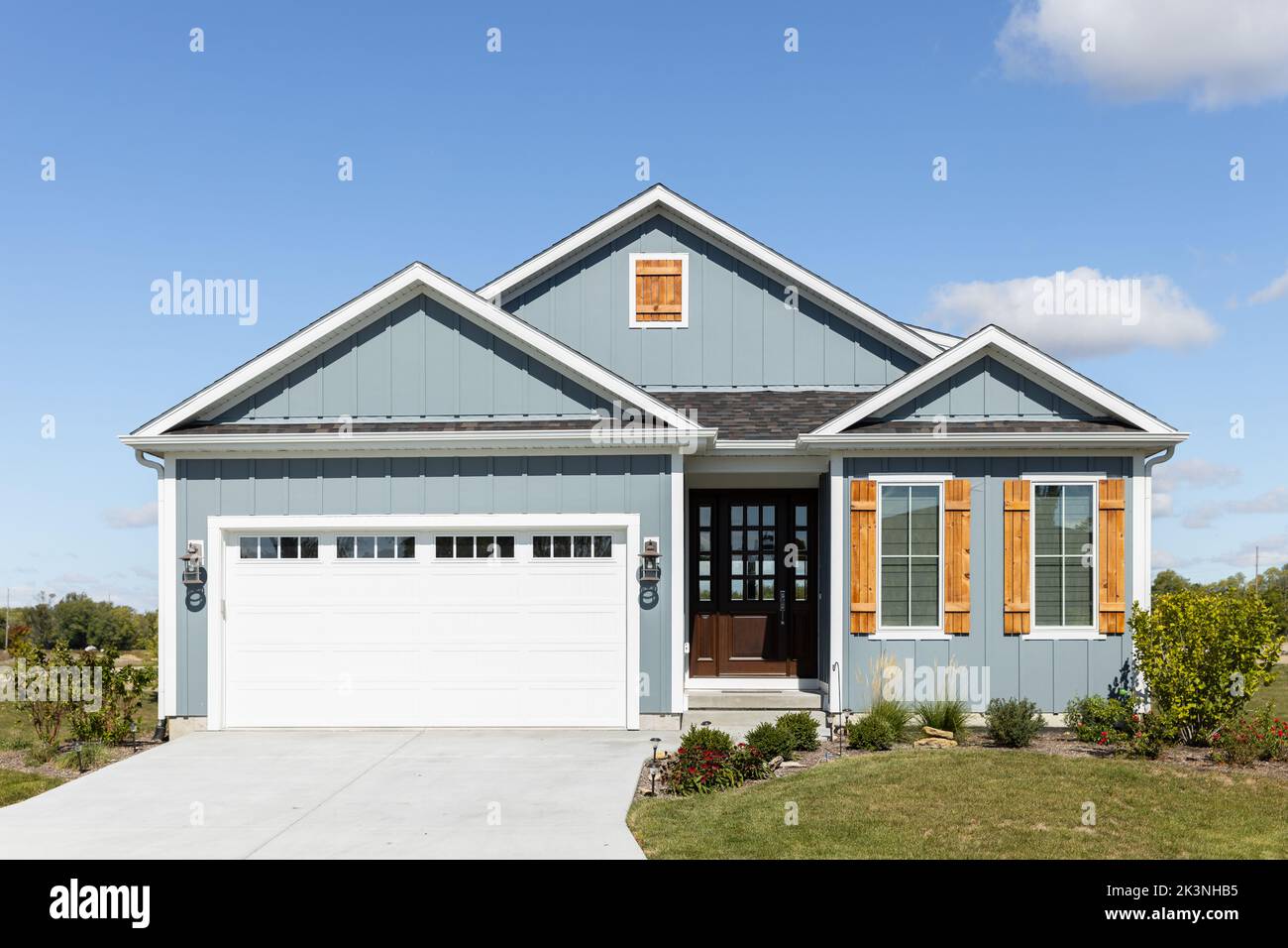 A light blue ranch home with a two car garage, wood window shutters,  vertical siding, and a dark wood front door. Stock Photo