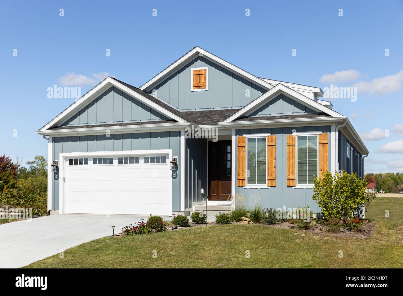 A light blue ranch home with a two car garage, wood window shutters,  vertical siding, and a dark wood front door. Stock Photo