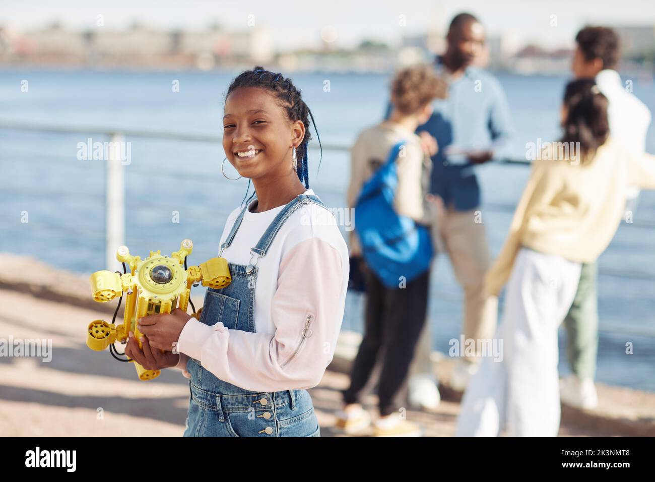 Waist up portrait of black teenage girl holding robot model and smiling at camera during outdoor engineering class with kids in background, copy space Stock Photo