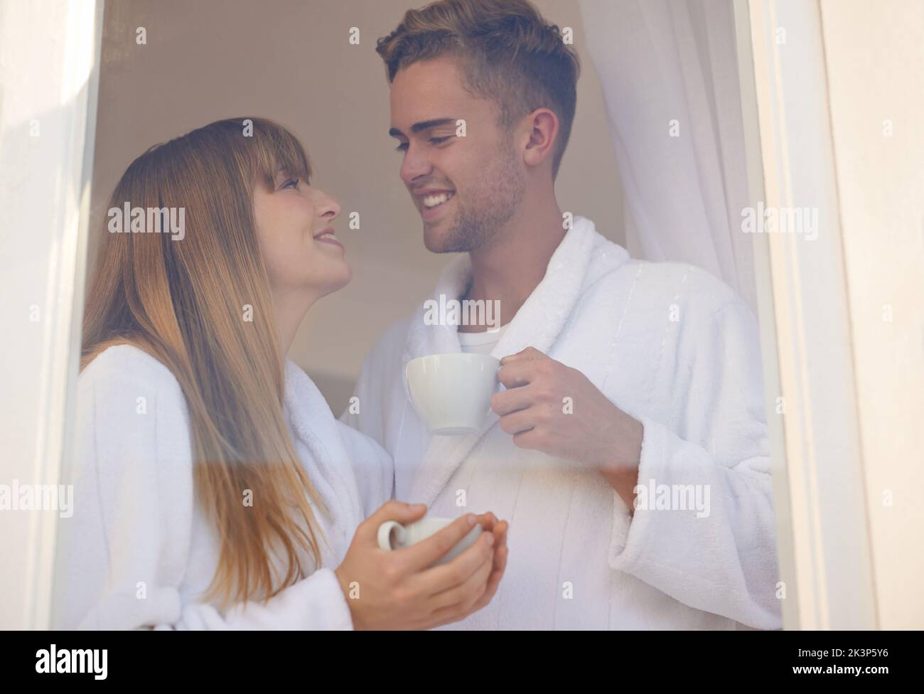 Starting the day together. A young couple drinking coffee while standing at a window wearing bathrobes. Stock Photo