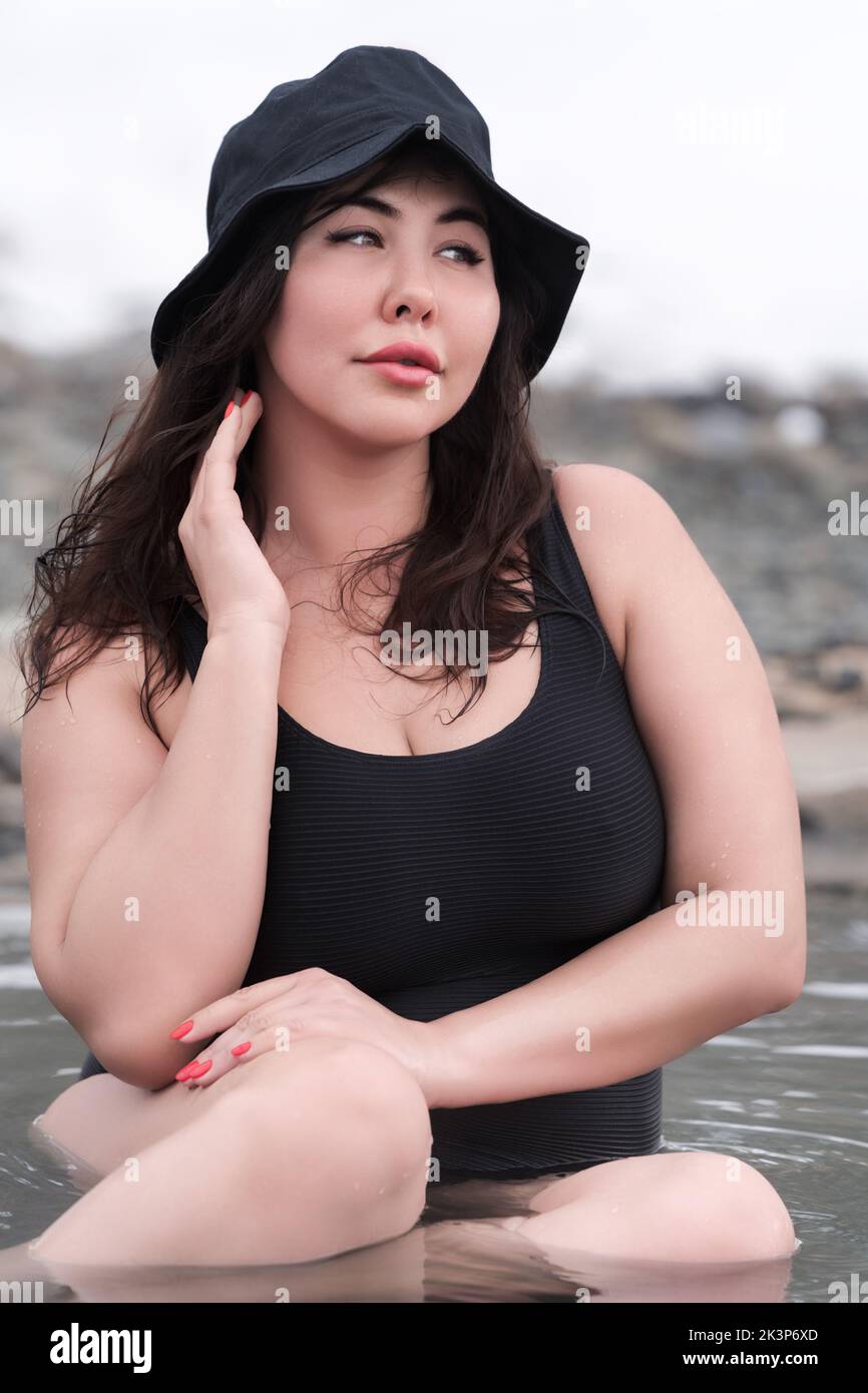 Front view of full figured young adult model in black one piece bathing swimsuit and panama hat sitting in outdoors pool at spa resort Stock Photo