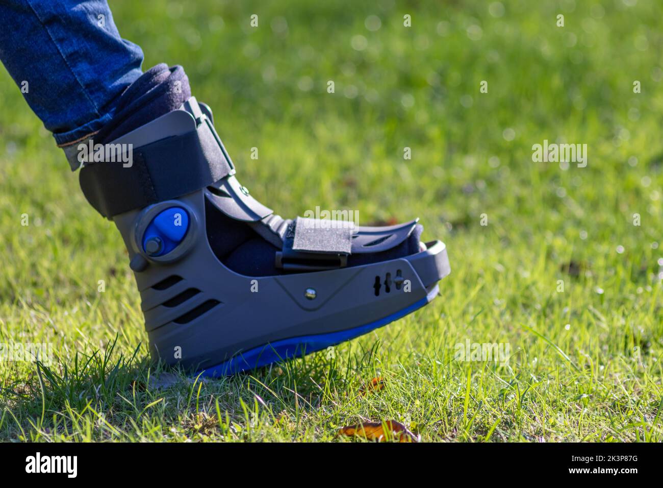 Boy with broken foot and orthopedic shoe or walker after bone fracture rests in public park on bench in green grass to recreate and rehabilitate Stock Photo