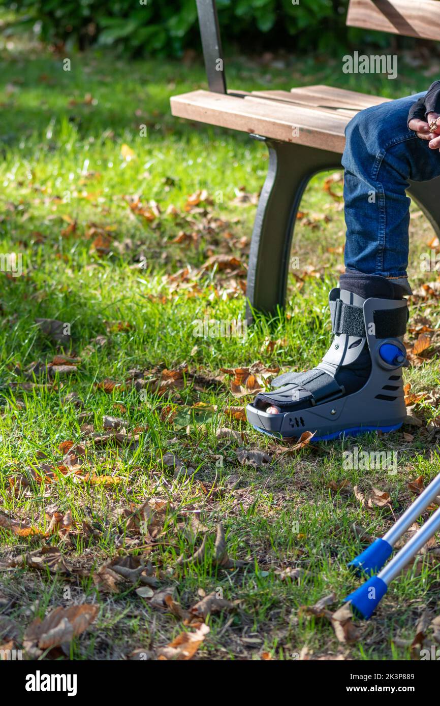 Boy with broken foot and orthopedic shoe or walker after bone fracture rests in public park on bench in green grass to recreate and rehabilitate Stock Photo