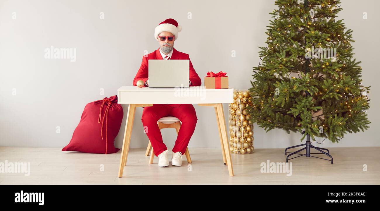 Funny modern Santa working on laptop while sitting at desk in office with Christmas tree Stock Photo