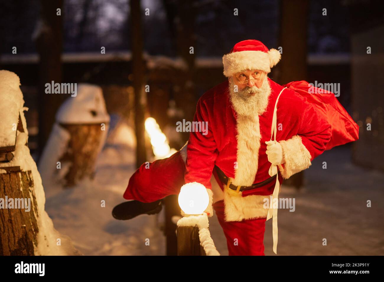 Portrait of traditional Santa Claus with sack of presents climbing over fence outdoors on Christmas eve Stock Photo