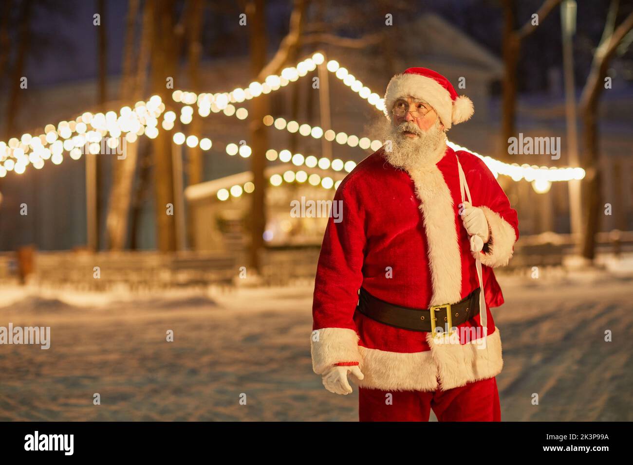 Waist up portrait of traditional Santa Claus carrying bag with presents at night with fairy lights in background, copy space Stock Photo