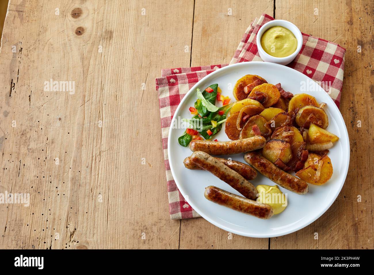 Top view of plate of bratwurst and bratkartoffeln with salad served on plate near bowl of mustard sauce and checkered napkin on wooden table Stock Photo