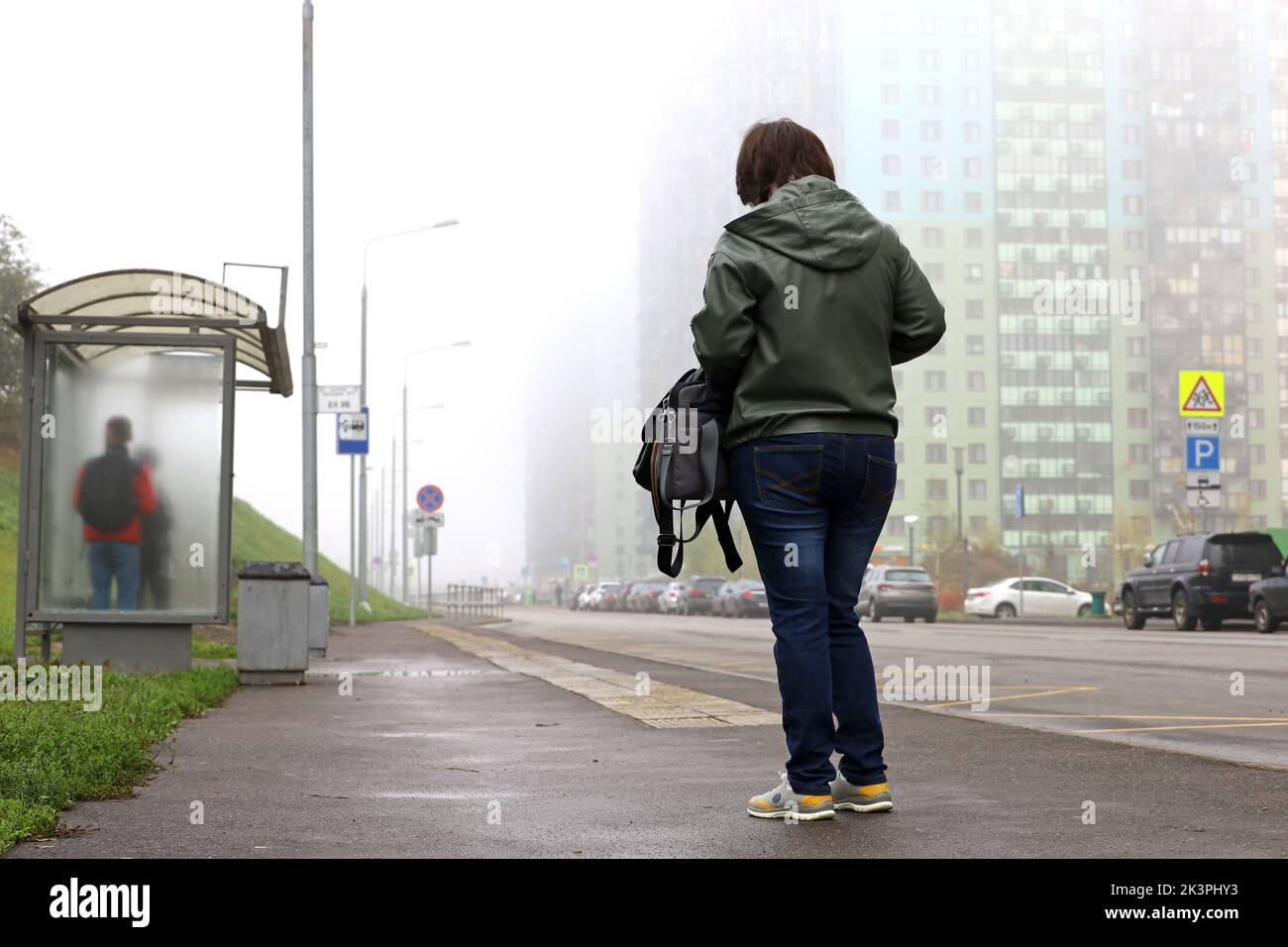 Girl standing at the bus stop on background of residential buildings in fog, public transport in autumn city Stock Photo