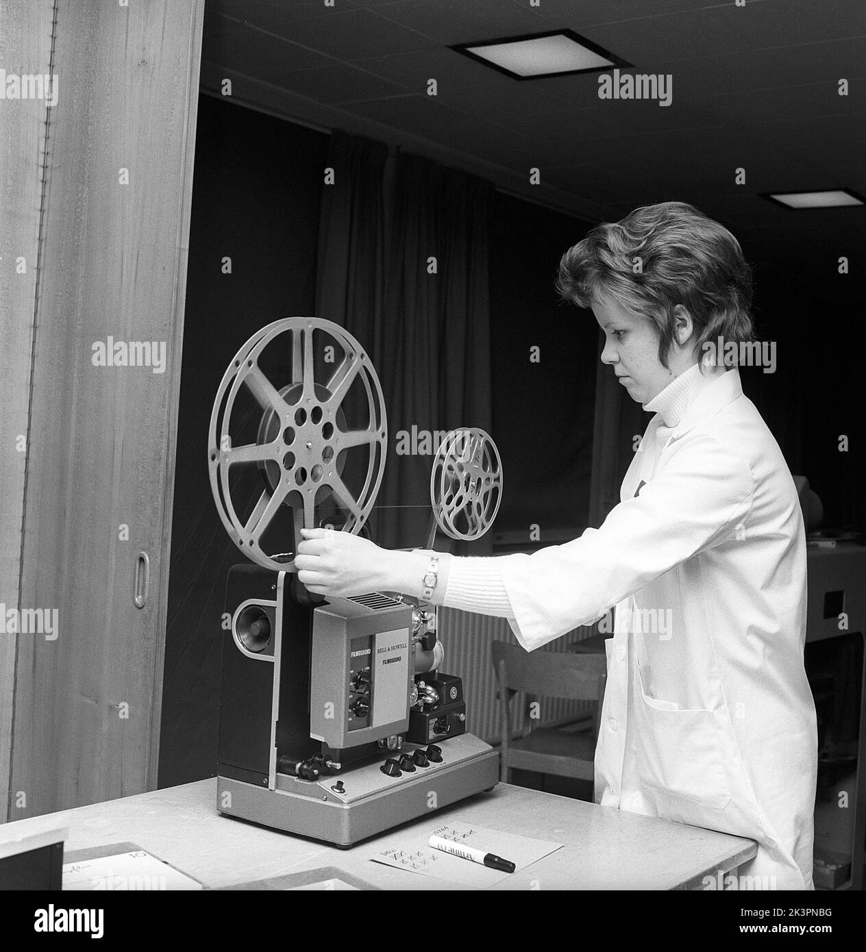 In the 1960s. A young female teacher with a film projector, mounting the film roll. Sometimes short films were shown in the classroom or in a special viewing room, not only for educational puroposes but for entertainment. The film had sound. Once the film roll was viewed, the film was rolled onto a reciever roll and needed to be winded back. Sweden 1968 ref CV77 Stock Photo