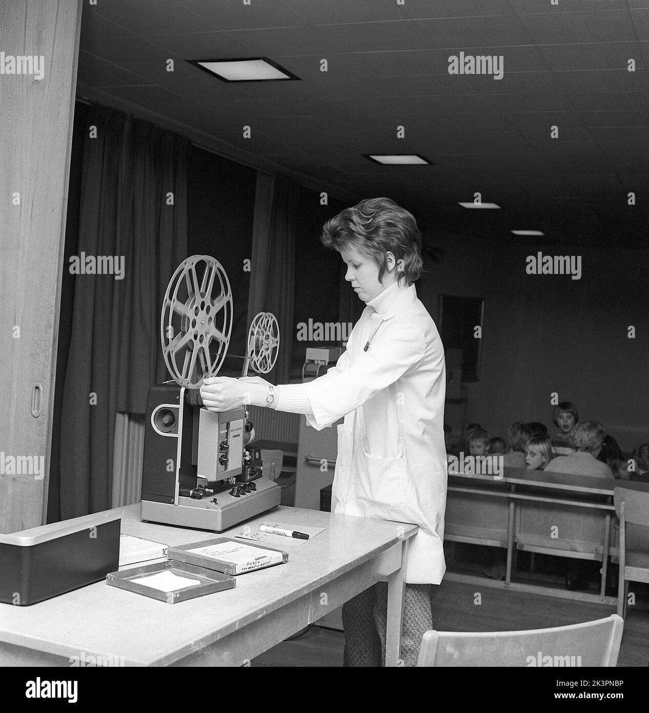 In the 1960s. A young female teacher with a film projector, mounting the film roll in the back of the classroom. Sometimes short films were shown in the classroom or in a special viewing room, not only for educational puroposes but for entertainment. The film had sound. Once the film roll was viewed, the film was rolled onto a reciever roll and needed to be winded back. The students waiting for the film to start is seen sitting in the background. Sweden 1968 ref CV77 Stock Photo