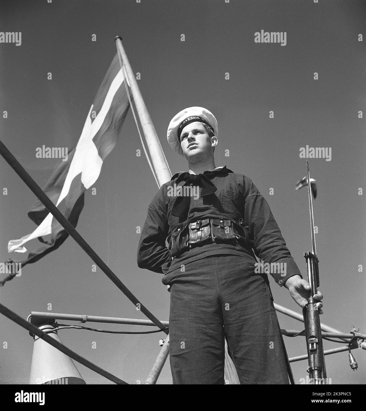 During World war II. The war ship Sverige during navy exercises at sea. Detail of a sailor standing guard on the ship with a rifle in his hand. The swedish naval flag is seen in the background. Sweden june 1940. Kristoffersson ref 141 Stock Photo