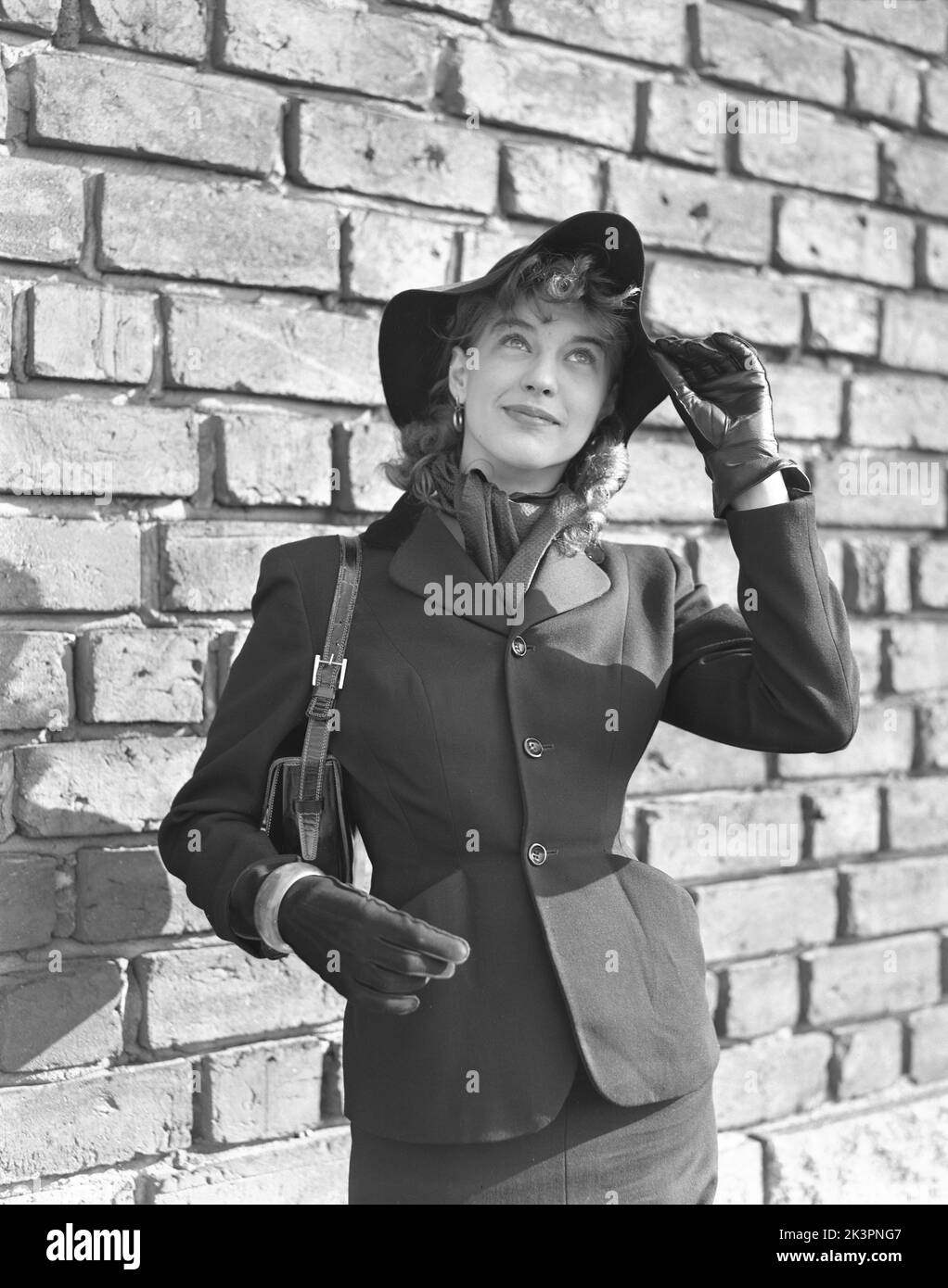 Fashionable in the 1950s. A young woman dressed in a jacket with a visible thin waistline, buttoned with three butoons. She wears a matching hat and and a scarf around her neck, black leather gloves and a handbag. Sweden 1952 Kristoffersson Ref 38K-20 Stock Photo