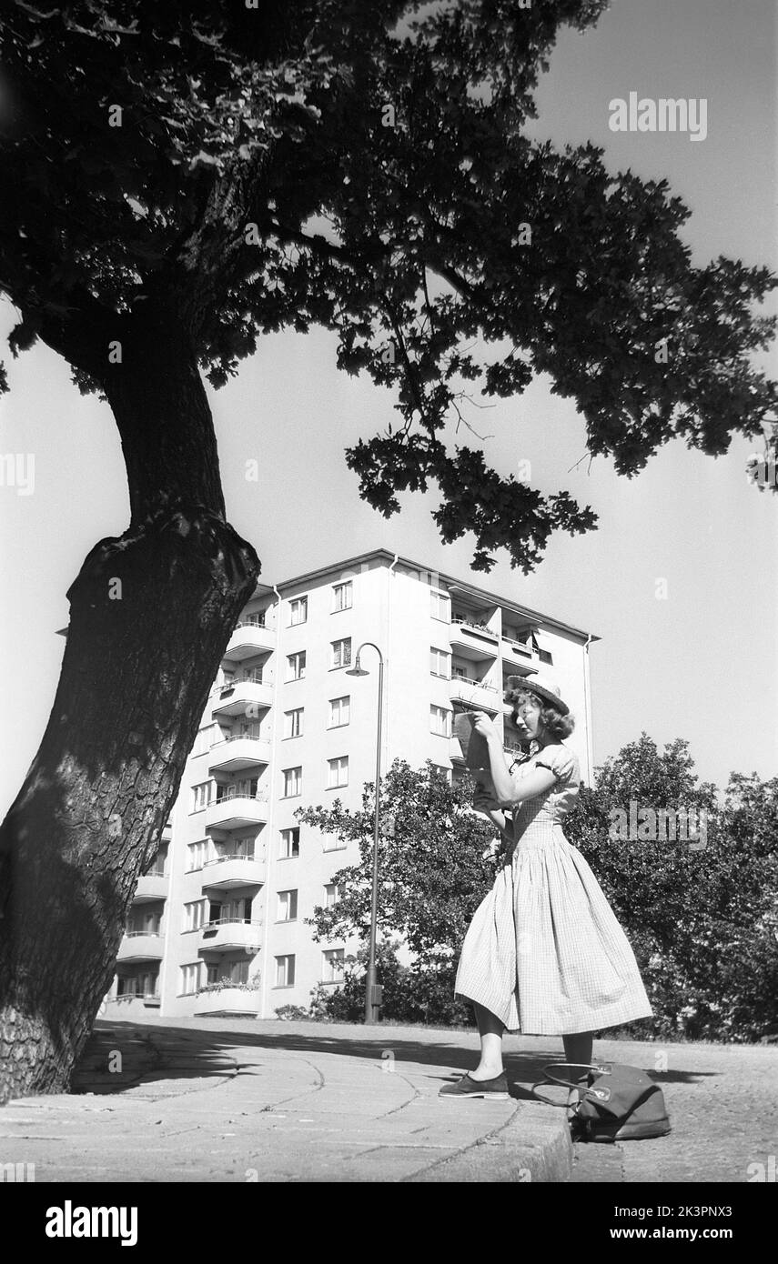 In the 1940s. A young woman is visiting Swedens captial city Stockholm and is pictured when standing in front of a large tree. She is wearing a summer dress and hat. It looks as if she is looking at something a map perhaps of what to see. The photograph has a composition that attracts. Spiky leaves, straight geometric lines in the building behind and the round forms of the dress. Stockholm Sweden 1949. Kristoffersson ref AO30-1 Stock Photo