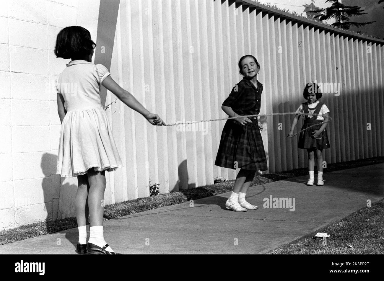 Mexico in the 1960s. Three girls are playing in the street, with a skipping rope - jumping rope. One girl in each end of the rope swinging it and a girl in the middle jumping over it. Mexico 1964. Roland Palm ref folder 2 Stock Photo