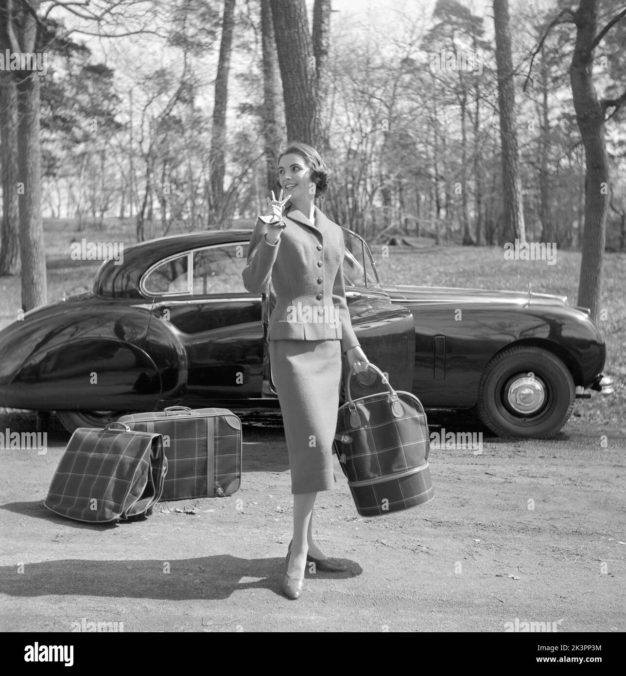 In the 1950s. A young woman in a matching outfit of jacket and skirt, typically 1950s. She stands in front of a beautifully curvy car with three bags and luggage in matching pattern. Sweden 1956 Stock Photo