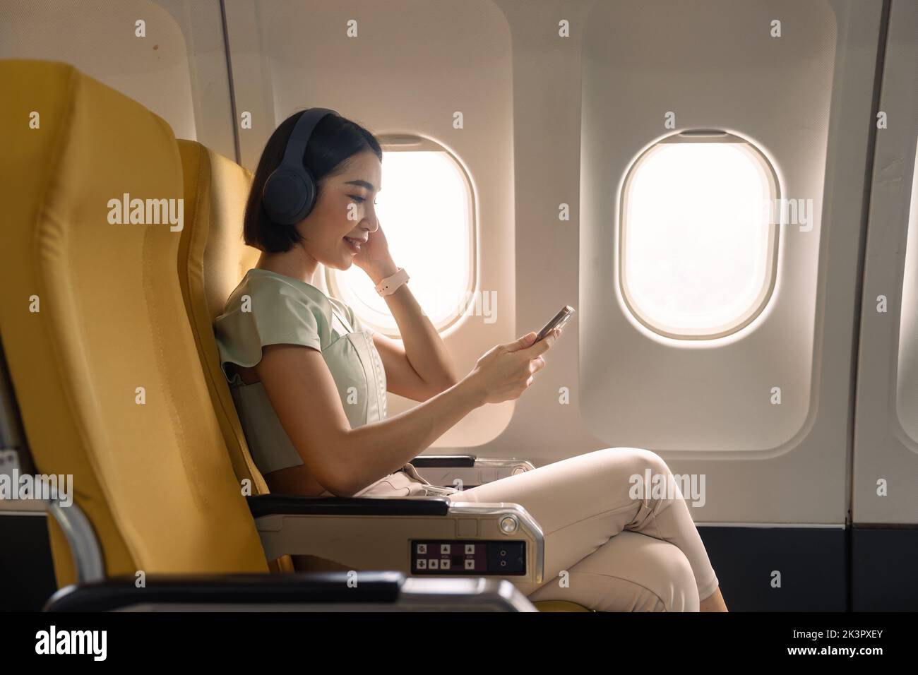 Young woman with mobile phone and headphones listening to music in airplane during flight Stock Photo
