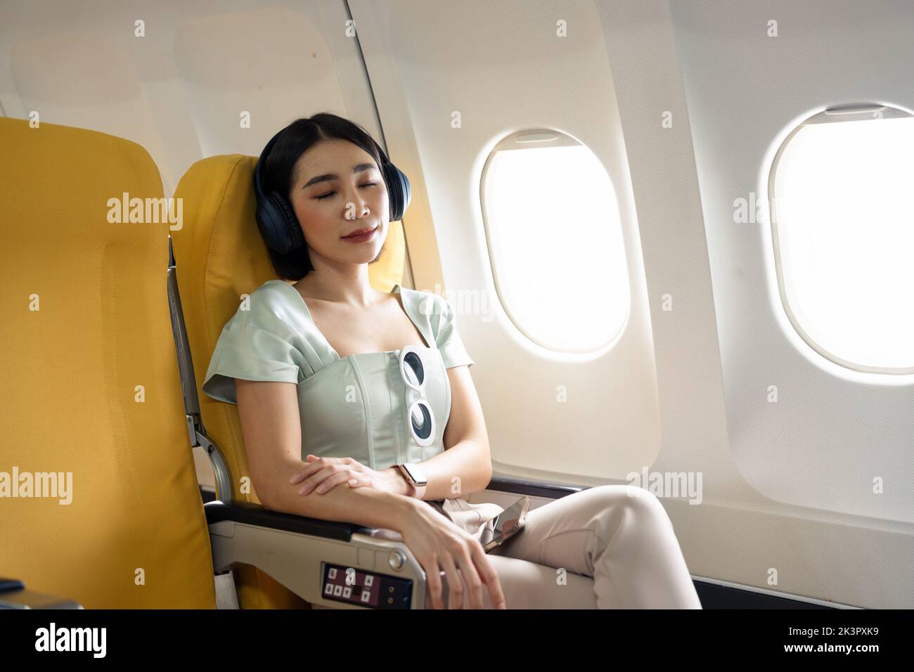 Tired asian lady napping with headphones on seat while traveling by airplane. Commercial transportation by planes. Stock Photo