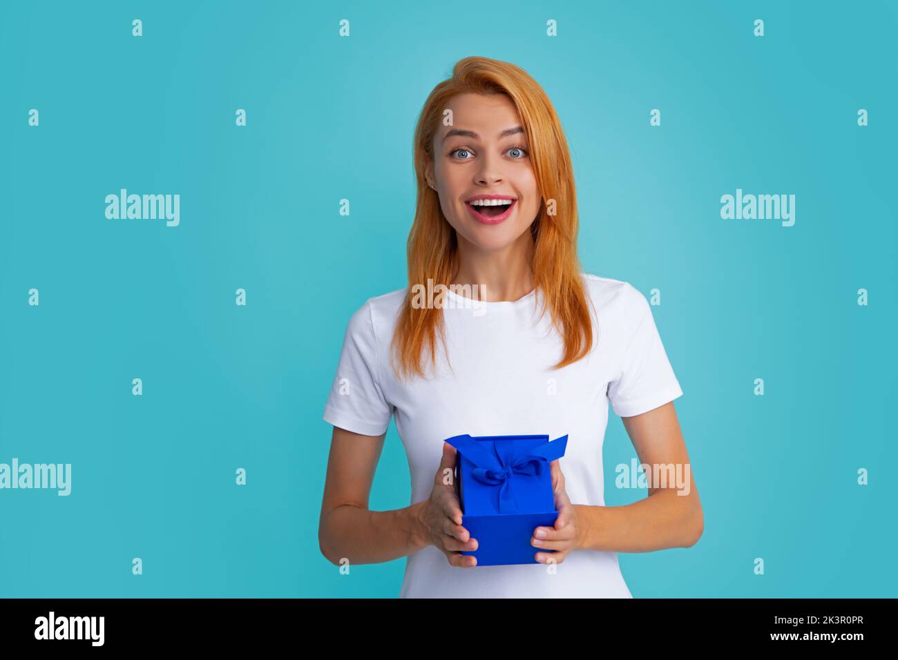 Surprised woman holding gift. Smiling beautiful young redhead woman in shirt hold present box with gift ribbon bow isolated on bright blue background Stock Photo