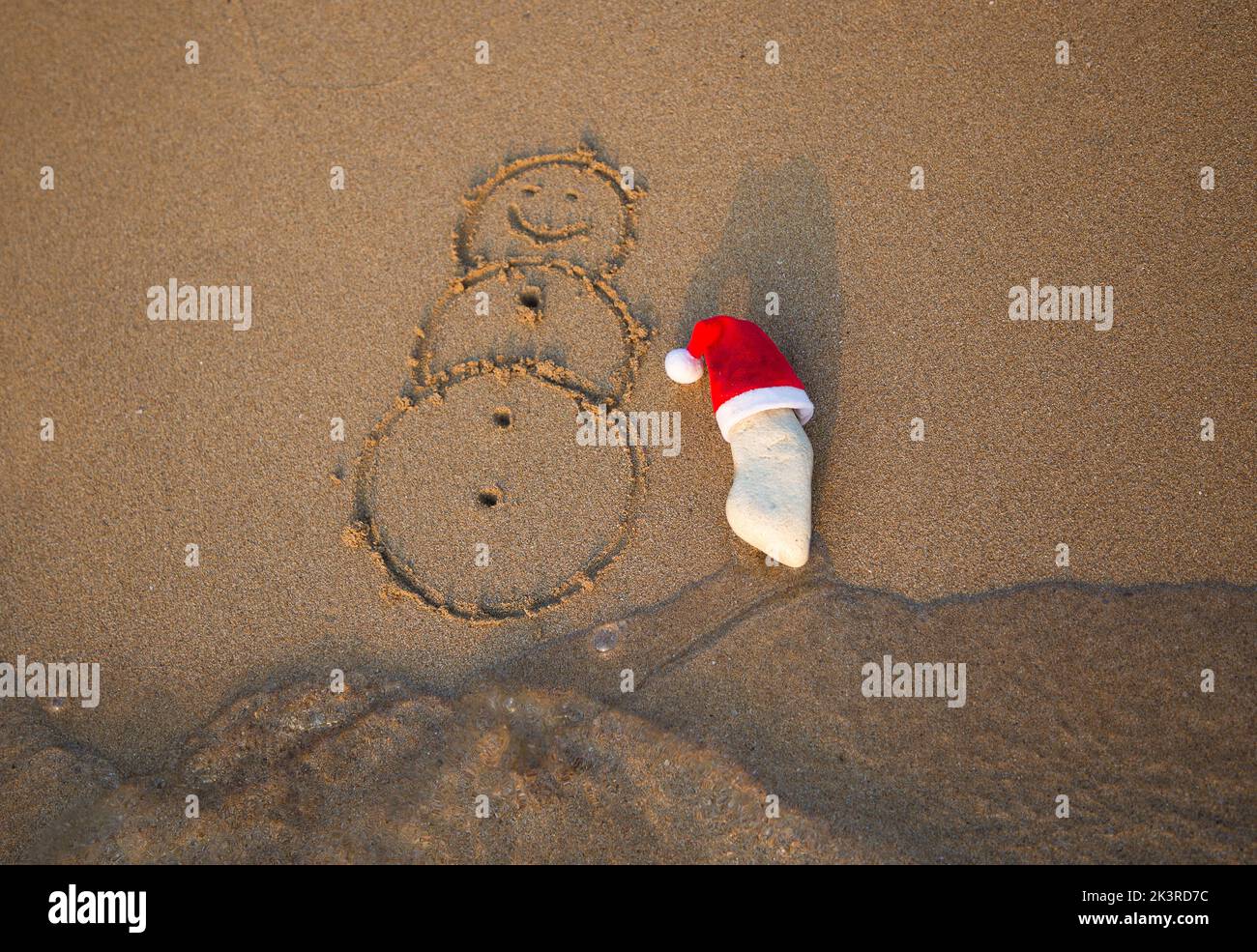 red Santa Claus hat on a white stone and a snowman painted on sand on coastal beach. The concept of celebrating Christmas holidays while traveling to Stock Photo