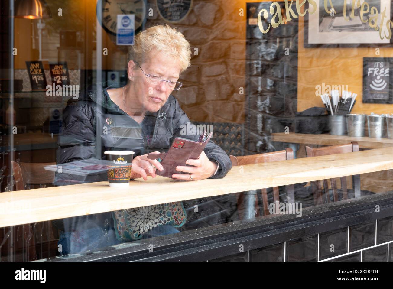 A cafe customer, with a coffee,  checks her phone whilst sitting at a window table inside the premises. Stock Photo