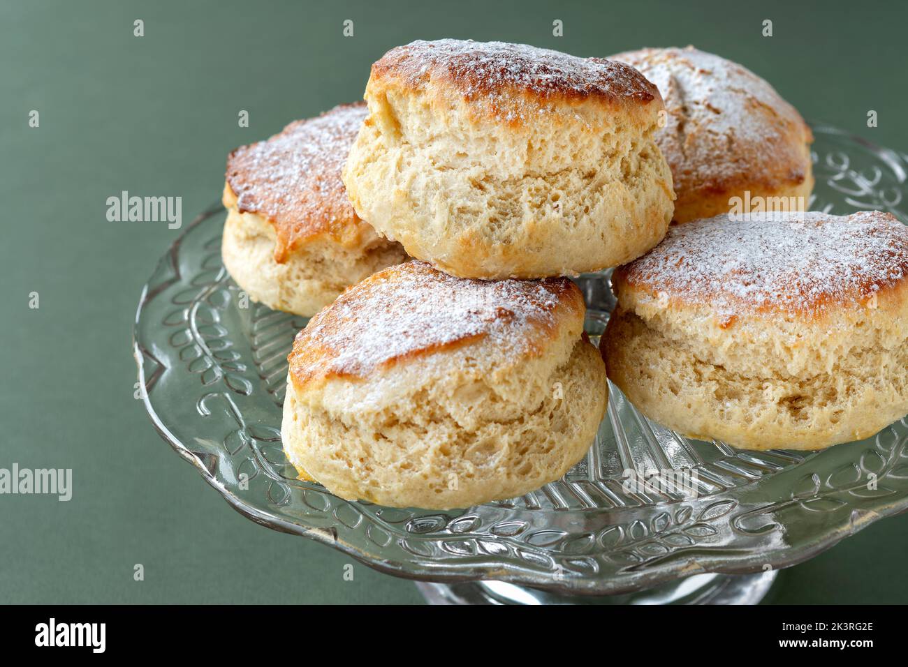 Freshly made traditional plain scones are shown on a glass cake stand. The cakes are lightly dusted with icing sugar and shown close up Stock Photo