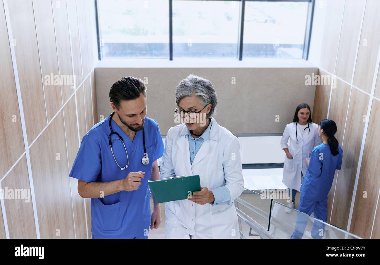 Mature doctor and male nurse reviewing patient's medical chart standing on stairs in hospital. Healthcare workers Stock Photo
