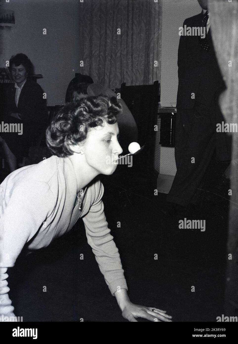 1956, historical, post-war Britain, inside a front room, a lady taking part in a party game, on the floor on her hands and knees with a spoon in her mouth balancing a small ball, as he she attempts to move it, England, UK. Stock Photo