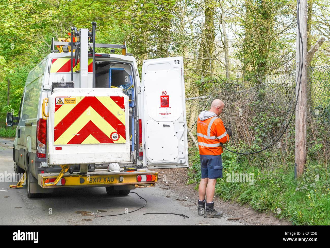 Rear view of BT Openreach vehicle, door open, and male worker fixing cable to a post in a UK country lane. Stock Photo