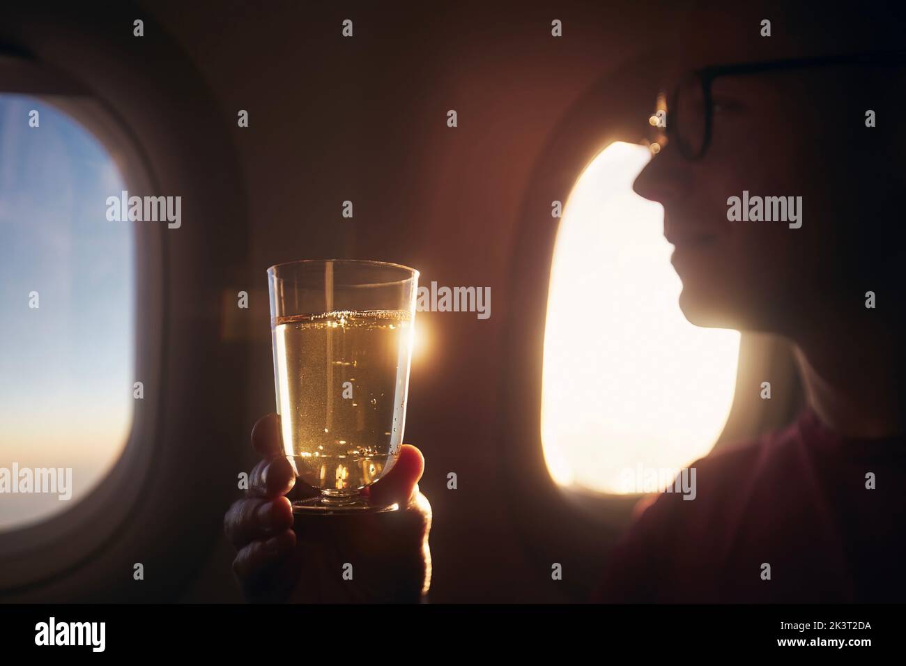Man enjoying drink during flight. Passenger holding glass of sparkling wine against airplane window at sunset. Stock Photo