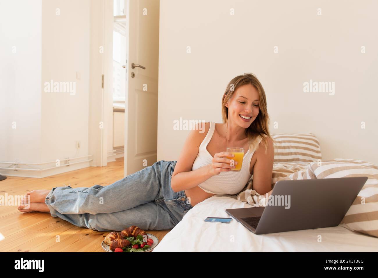 Cheerful young woman holding orange juice near laptop and breakfast in bedroom,stock image Stock Photo