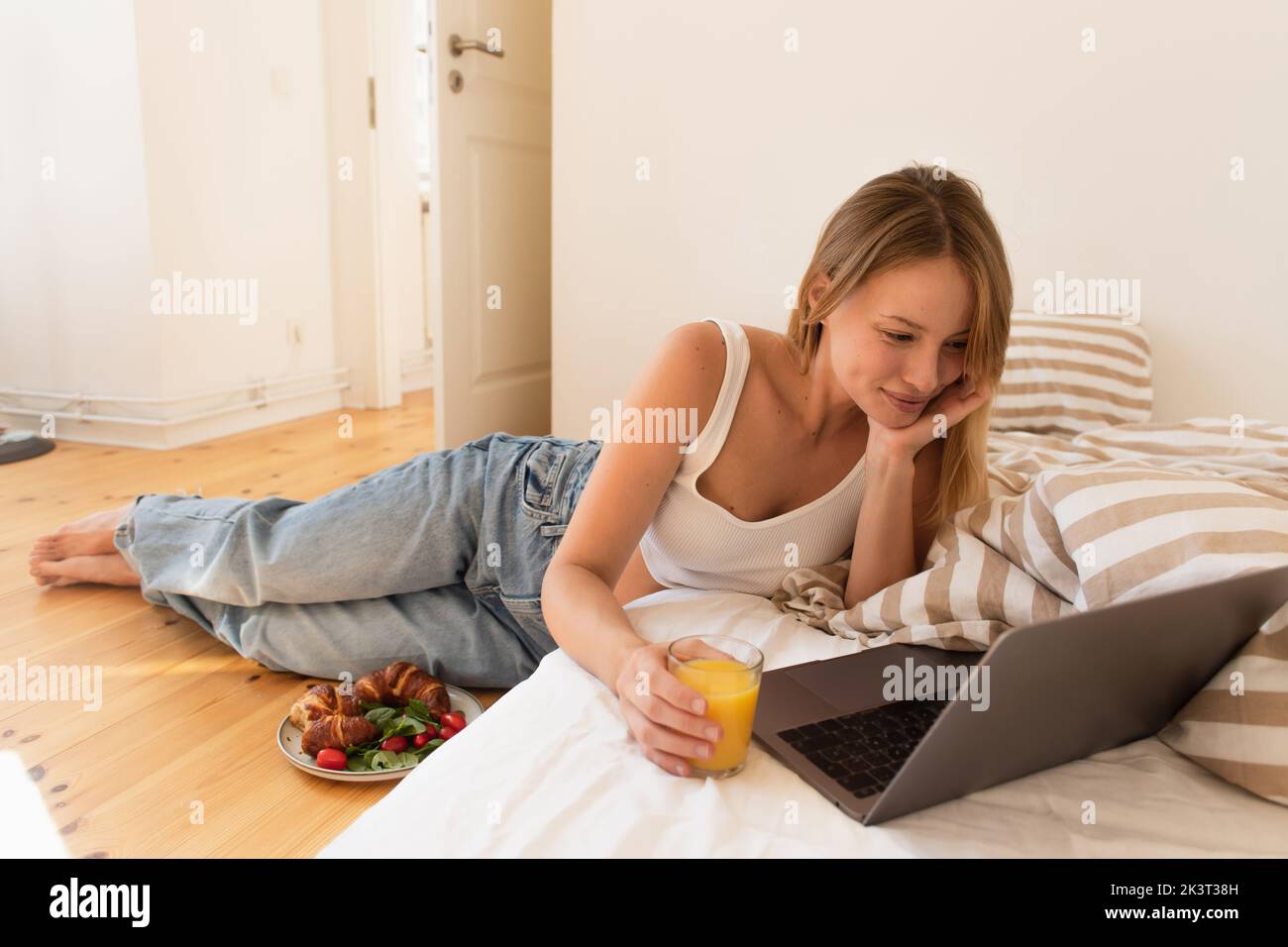 Smiling freelancer looking at laptop and holding orange juice near tasty breakfast on floor at home,stock image Stock Photo