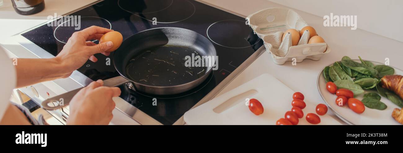 Cropped view of woman holding egg near frying pan and cherry tomatoes in kitchen, banner,stock image Stock Photo