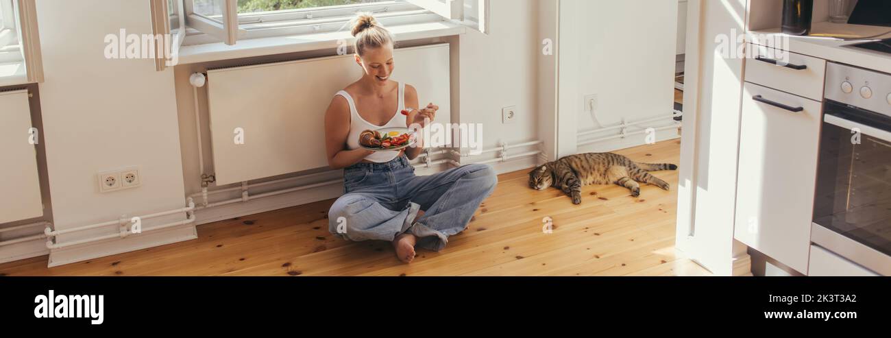 Cheerful woman holding plate with delicious breakfast near scottish fold cat on floor at home, banner,stock image Stock Photo