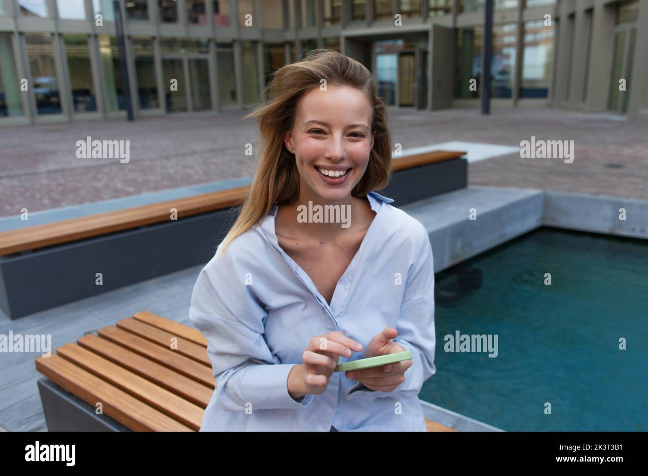 Cheerful blonde woman in shirt holding smartphone and looking at camera on bench on street,stock image Stock Photo