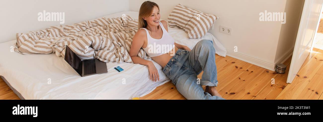 Cheerful woman sitting near laptop and credit card on bed, banner,stock image Stock Photo
