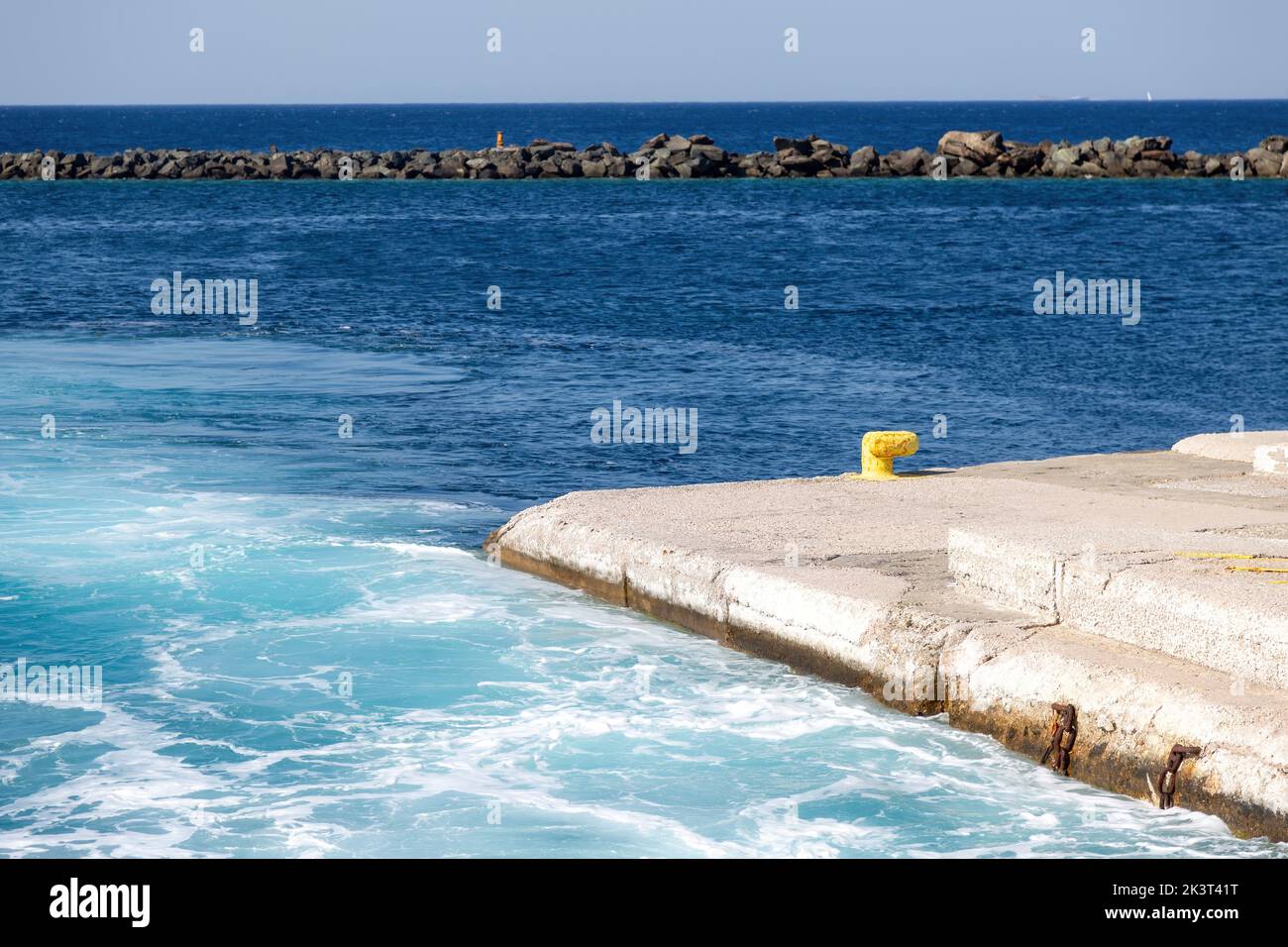 Greece, Naxos harbor destination Cyclades islands. View of corner of jetty and stone breakwater. Calm sea with foam at dock, sunny day, blue sky backg Stock Photo