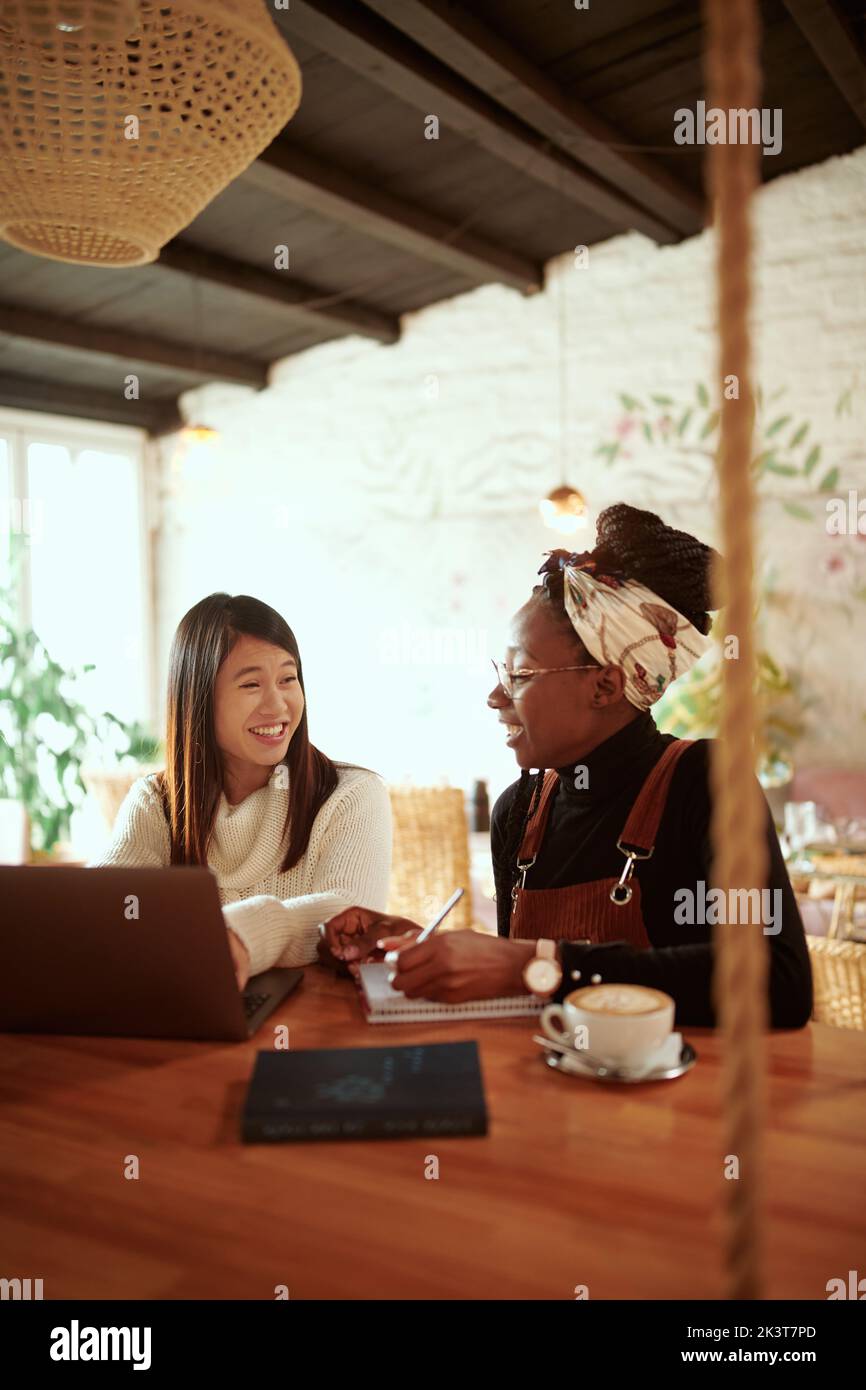 Two beautiful multicultural female students sitting in a cafe and following online lecture on the laptop. Technology in education. Stock Photo