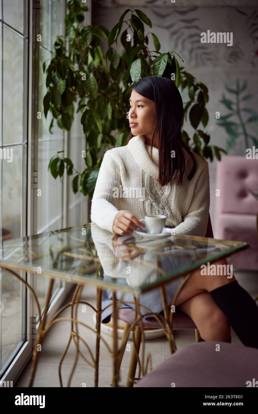A thoughtful asian woman is sitting in a coffee shop and looking trough ...
