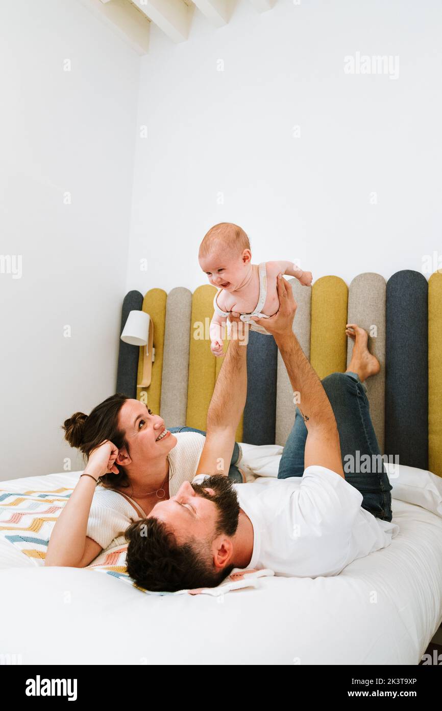 Father pretending that playful baby flying like superhero while chilling together with wife and kid on soft bed at home Stock Photo