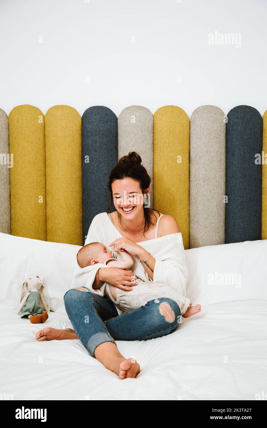 Young woman embracing with love and feeding infant while sitting on soft bed against multicolored headboard in light modern bedroom Stock Photo