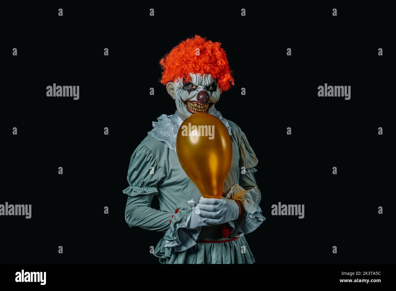 a red-hair creepy evil clown, staring at the observer while is holding a golden balloon in his hands, wearing white gloves, standing on a black backgr Stock Photo
