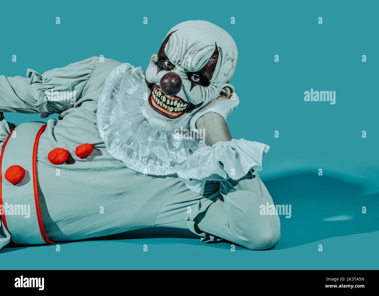 a creepy bald evil clown, with a menacing smile, wearing a gray costume with a white ruff, is lying on his side, leaning on one arm, on a blue backgro Stock Photo
